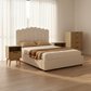 Beige upholstered bed with wooden nightstands and dresser in a bright bedroom.
