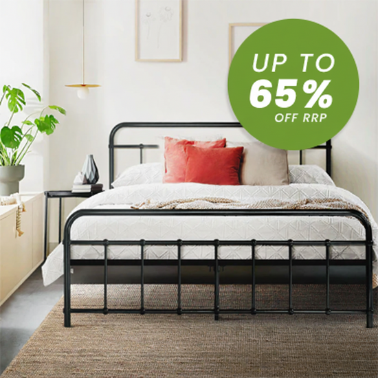 A black metal bed frame with a white quilted bedding set and two red accent pillows, shown in a bright bedroom setting with a green circular sale overlay reading 'UP TO 65% OFF RRP'.