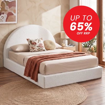 A low-profile upholstered bed with a rounded headboard in a warm beige fabric, styled with beige bedding, a rust-brown throw blanket, and decorative pillows. The bed is set in a cozy bedroom with a wooden floor, a round jute rug, and a red promotional sticker overlay that reads 'UP TO 65% OFF RRP'.